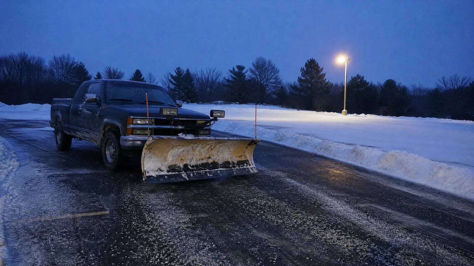 Pickup with a snow plow blade lowered on a partly cleared commercial parking lot before dawn